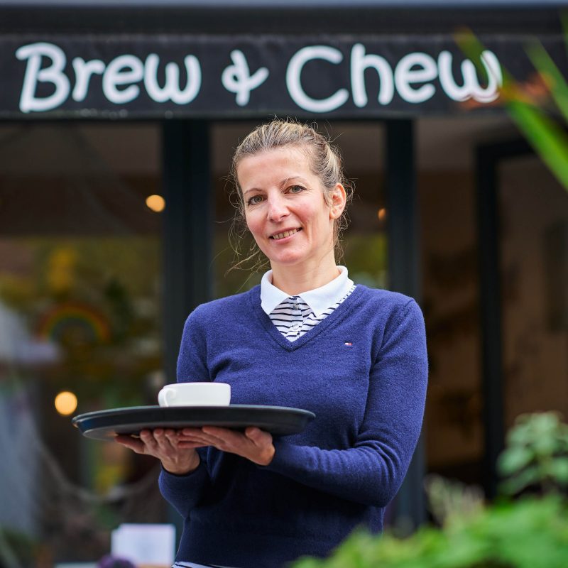A smiling woman wearing a blue jumper holds a tray carrying a white mug outside a cafe in Perth