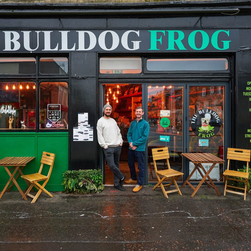Two smiling men stand in a seating area outside a cafe with black walls, windows, and a door with a cartoon logo printed on to it