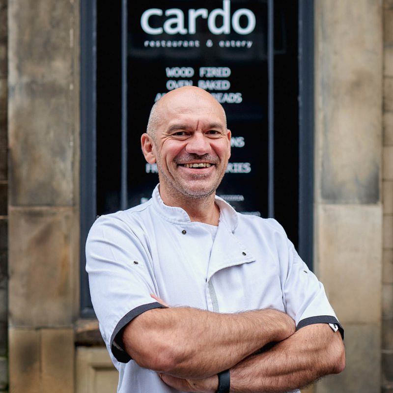 A smiling man wearing a white chef's uniform stands with his arms crossed outside a restaurant in Perth
