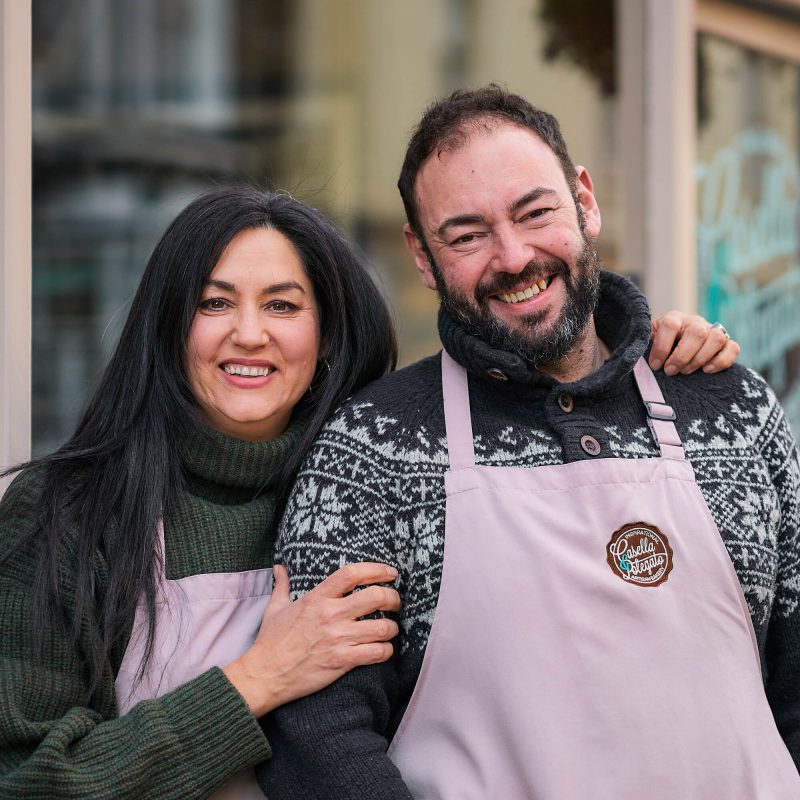 A smiling woman rests her hand on a smiling man's arm, both wearing matching pale pink aprons