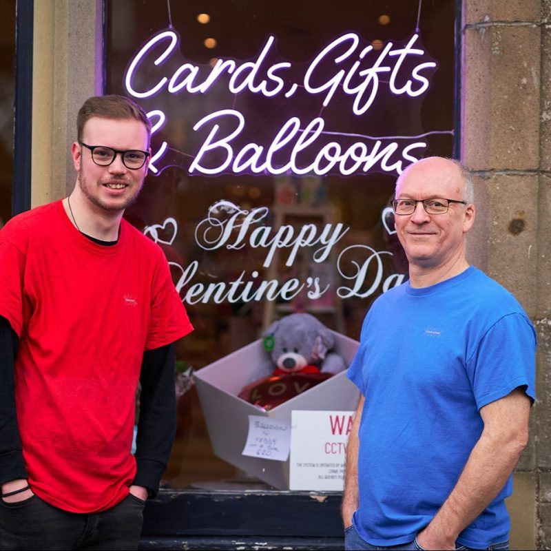 An older man wearing a blue shirt stands next to a younger man wearing a red shirt outside a large window with a pink neon sign