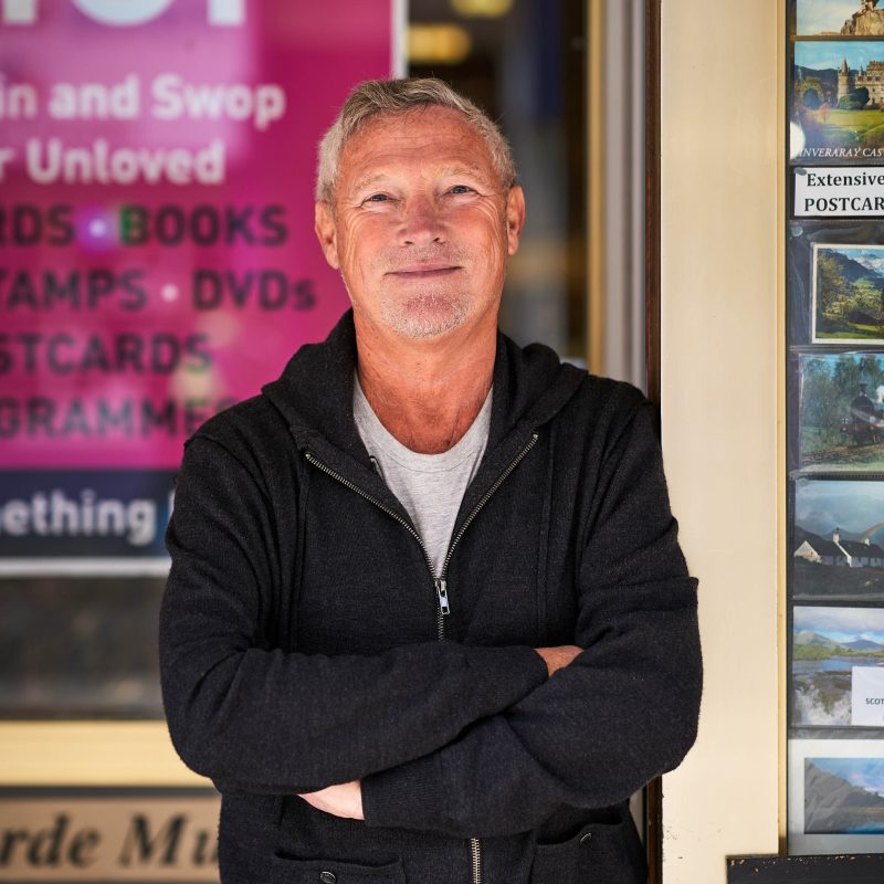 A man with grey hair wearing a black jumper stands with his arms crossed outside a window displaying a pink sign and postcards