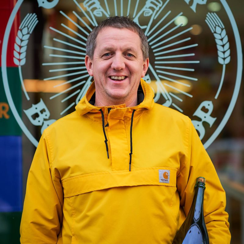 A smiling man wearing a yellow jacket holds a glass bottle while standing outside a brewery