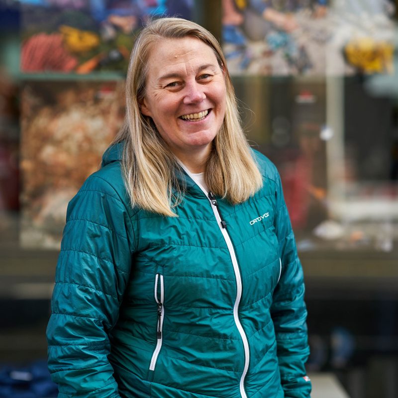 A smiling woman with blonde hair wearing a blue jacket stands outside a window with a colourful display