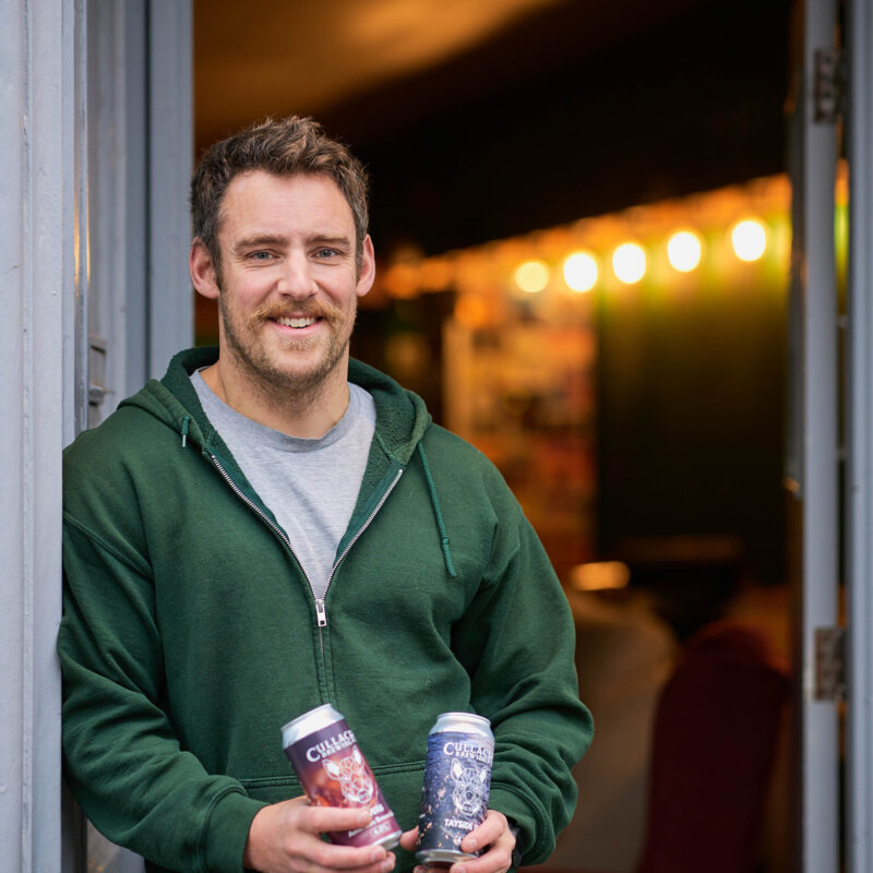 Smiling bearded man holding tins of beer, leaning against a door wearing a green hoodie and grey t shirt.