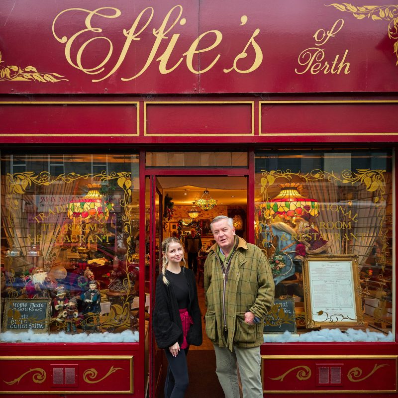 A young woman wearing black stands next to an older man wearing green outside a cafe painted red with two decorated windows