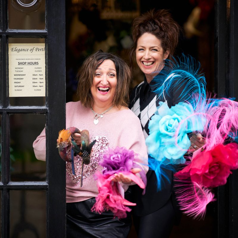 Two smiling women holding several brightly coloured hats with different patterns and textures stand in a dark doorway in Perth