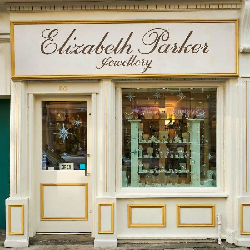 A white shop has a sign reading Elizabeth Parker Jewellery written in cursive above a window displaying an array of jewellery
