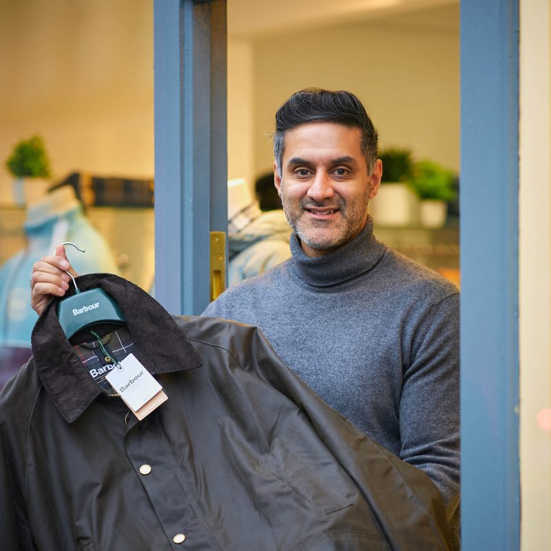 A smiling man wearing a grey shirt holds a black men's collared jacket in a doorway outside a shop