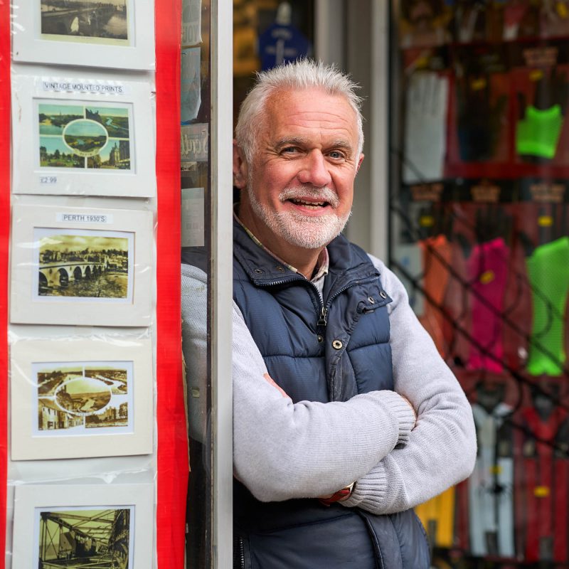 A smiling man with white hair stands with his arms crossed next to printed pictures in a doorway outside a shop