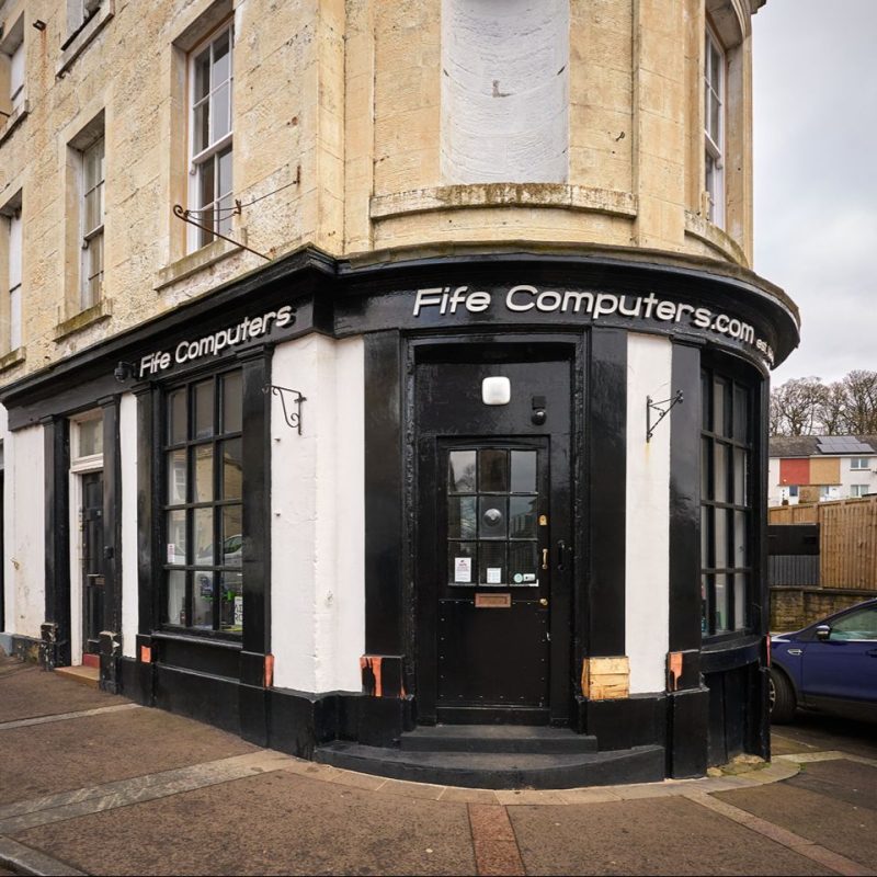 A curved shop front on a road corner with large black panel windows, a large black door and a black sign with white font