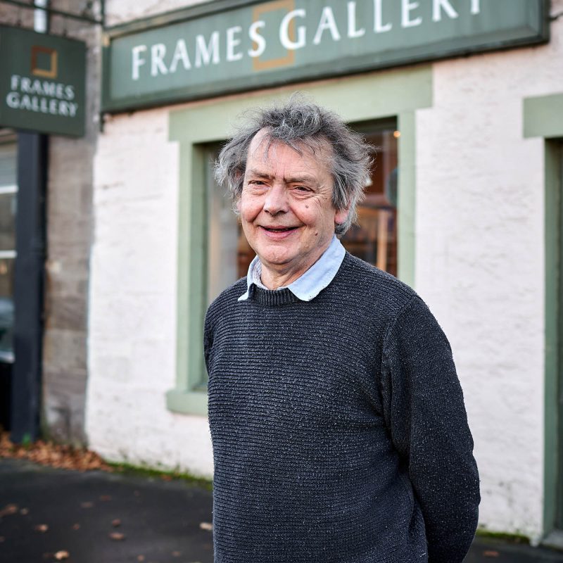 An older man with grey hair wearing a grey jumper stands outside a white shop with a large green sign