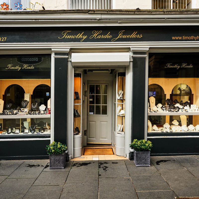 A black and white shop front with signs written in gold cursive font and two large windows displaying a collection of fine jewellery