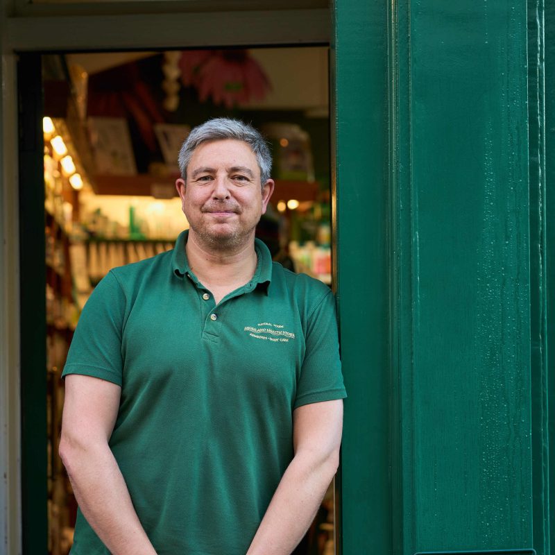 A man with grey hair wearing a green shirt leans against a doorway painted the same shade of green outside a shop
