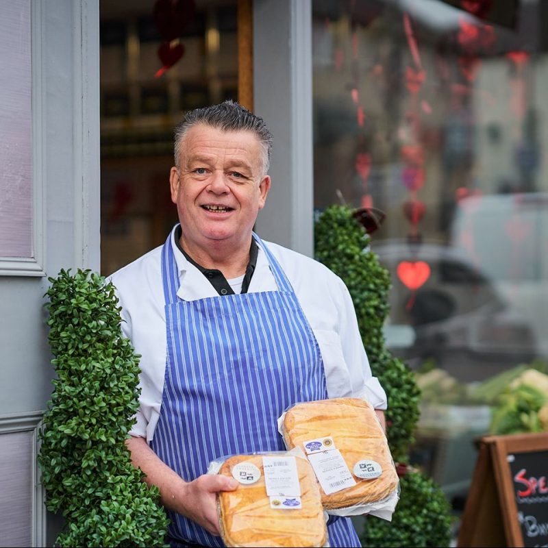 A smiling man wearing a stripped apron holds two packaged steak pies while standing in front of a doorway and window