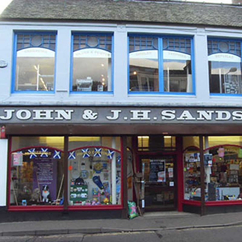 A shop front with very large windows with red frames displaying miscellaneous objects underneath large windows with blue frames.