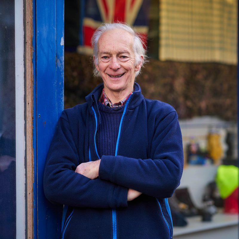 A smiling older man wearing a blue jumper has his arms crossed and stands in a doorway outside a shop