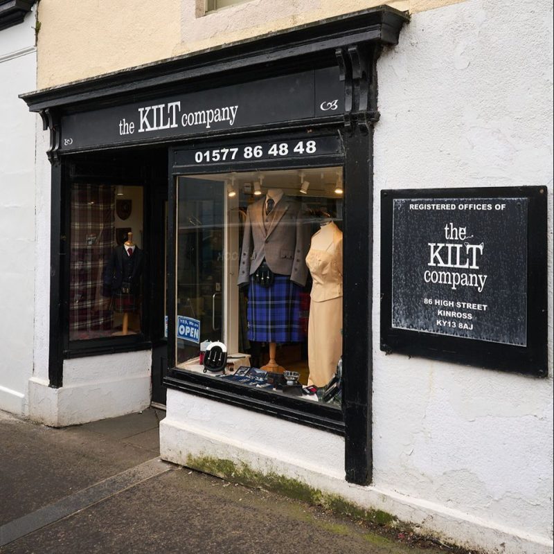 A black shop front with a black sign and a large window displaying mannequins wearing kilts and formal Scottish clothing
