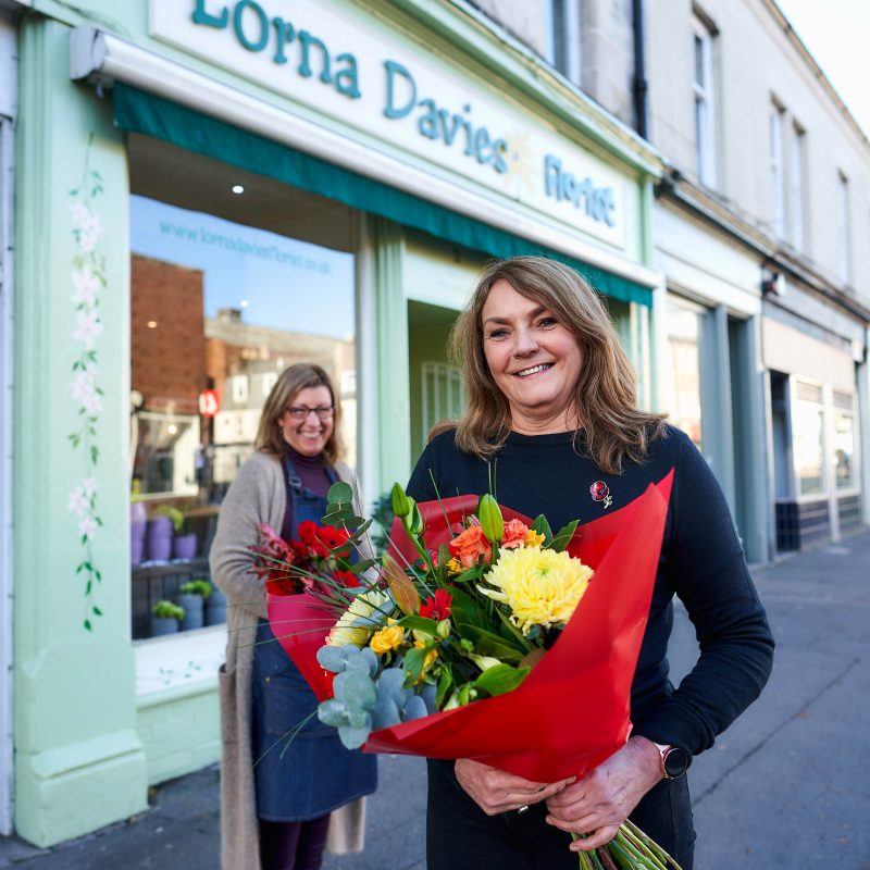 A smiling woman holding a bouquet of colourful flowers stands next to a smiling woman wearing an apron outside a green florist