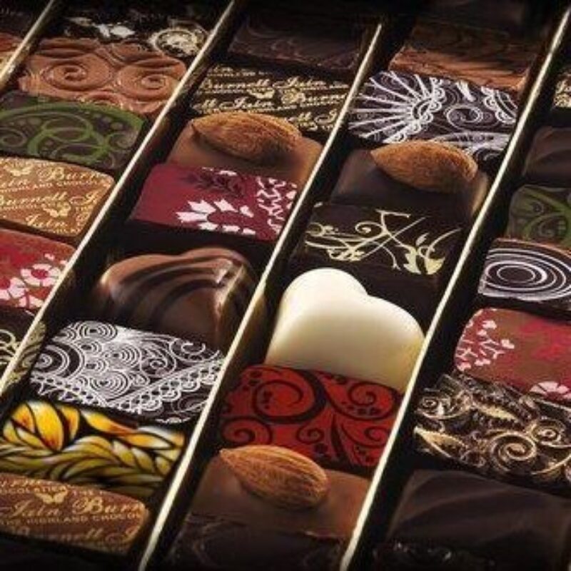 Two heart-shaped chocolates sit in a box amongst square chocolates with varying printed colourful designs