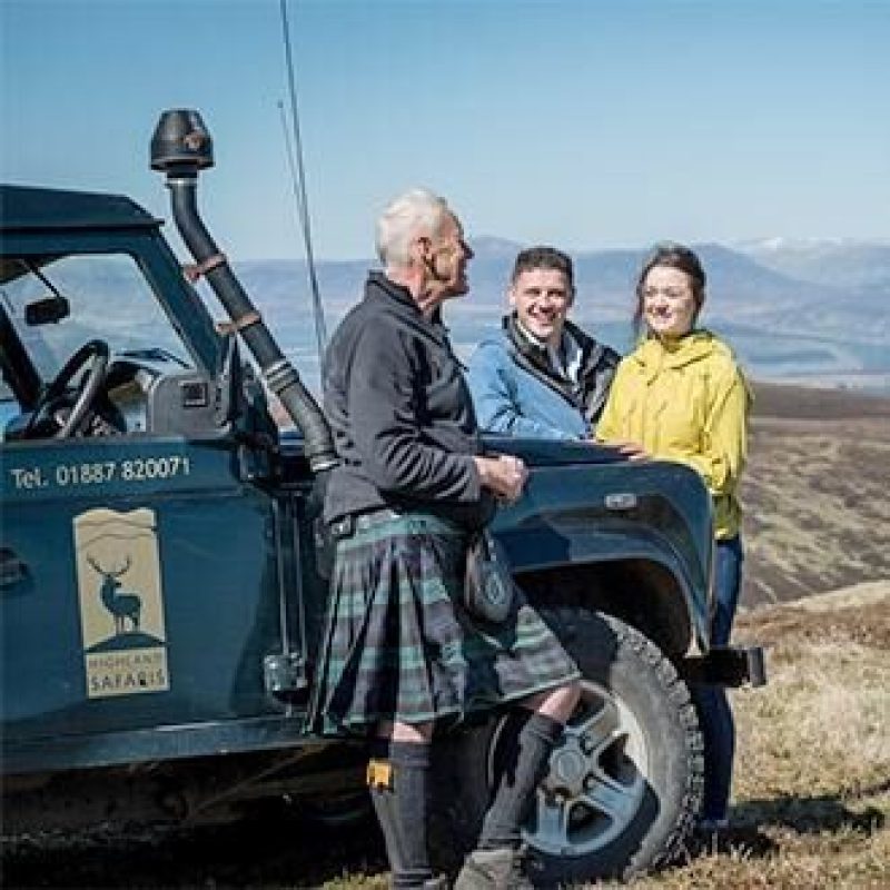 A man wearing a kilt stands next to a younger man and woman next to a green off road car in a mountainous landscape