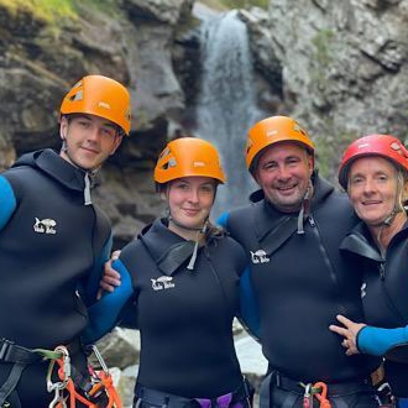 A group of four people wearing wet suits and helmets stand arm in arm in front of a large rocky waterfall