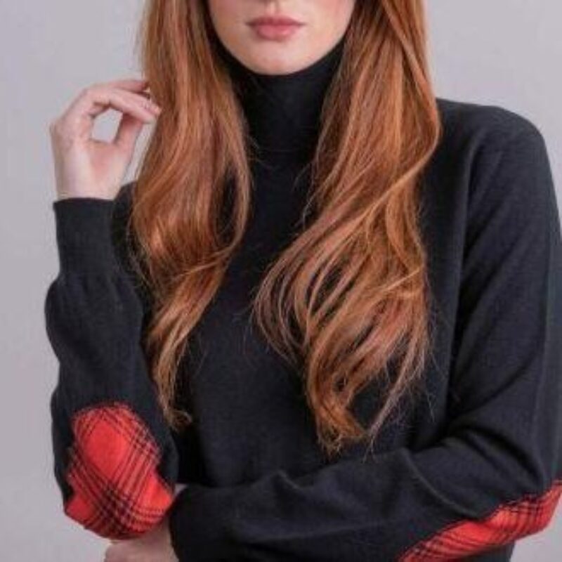 A woman with long ginger hair wearing a black sweater with red elbow patches poses in front of a grey background
