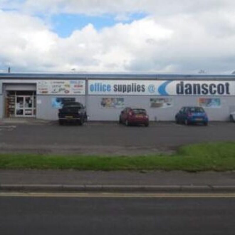 A short white building with a large white sign reading Danscot office supplies sits in a car park populated by four cars