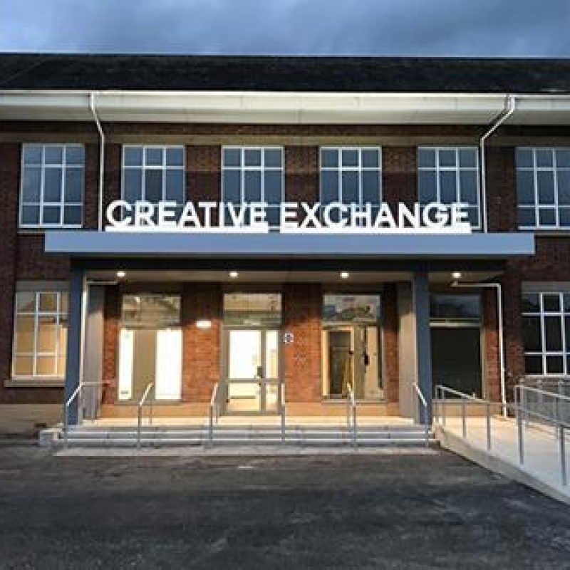 A large stone building with several windows has a large entry way with a white sign reading Creative Exchange above the doors