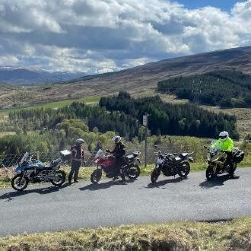 Five motorcycles are parked in a line in on a mountainous country road, with three riders gathered around them