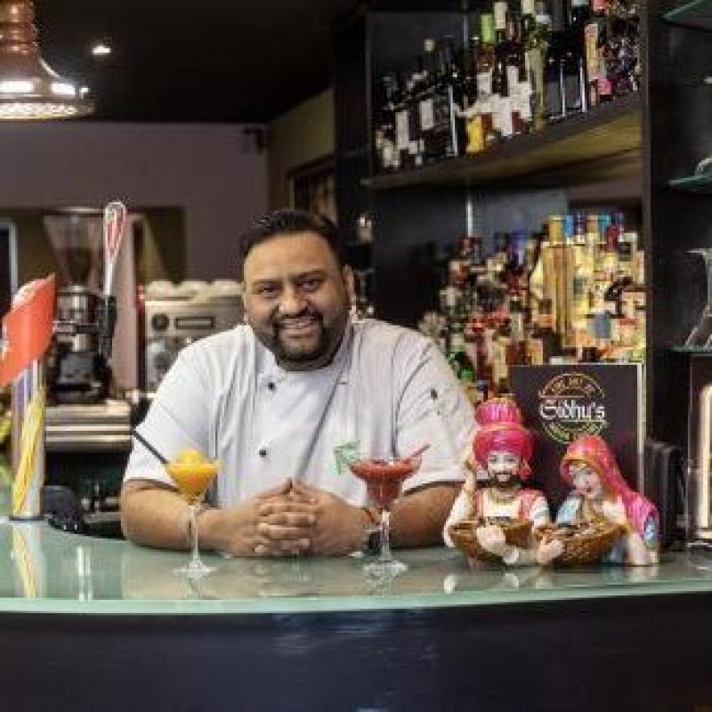 A smiling man wearing a white chef's uniform stands behind a bar with two colourful cocktails sitting next to two figurines