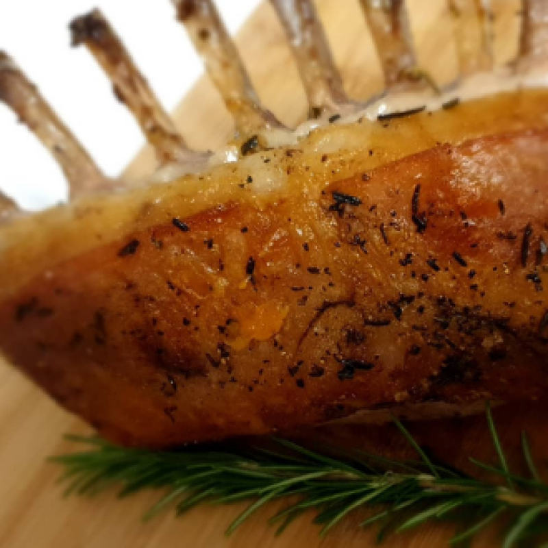 A roasted rack of seasoned lamb sits on a wooden chopping board next to a stick of rosemary