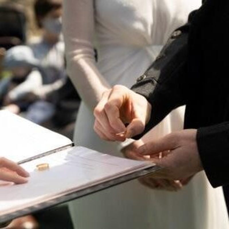 A groom standing next to a bride picks up a gold wedding band from a book being held by an officiator