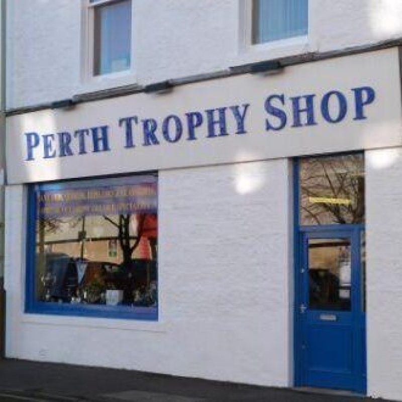 A large white sign with blue writing reading Perth Trophy Shop sits above a window displaying several different trophies