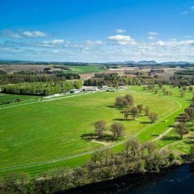 A horse racing track next to a river with a large green field populated with trees and rolling fields and mountains in the distance