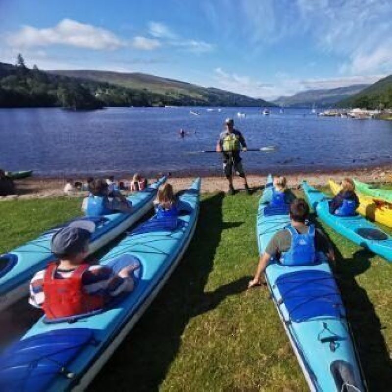 A group of people wearing life jackets sit in brightly coloured canoes at the edge of a loch and watch an instructor