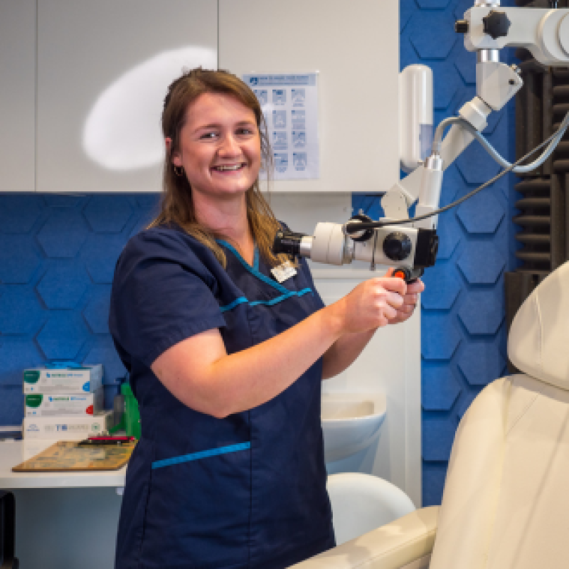 A smiling woman wearing black scrubs stands in a room and holds equipment next to a white chair