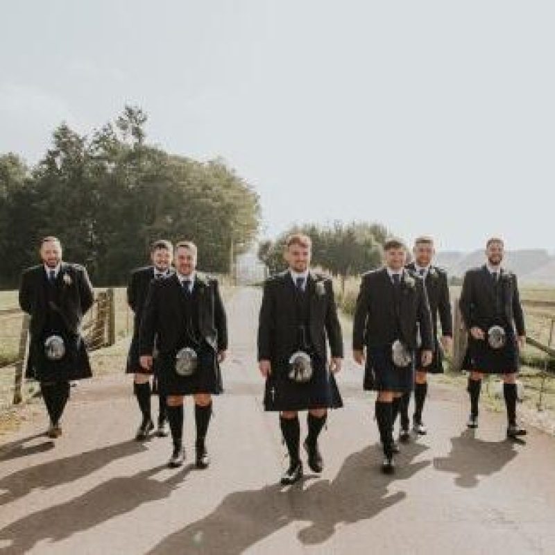A group of men wearing matching black blazers, kilts, and sporrans walk along a country road