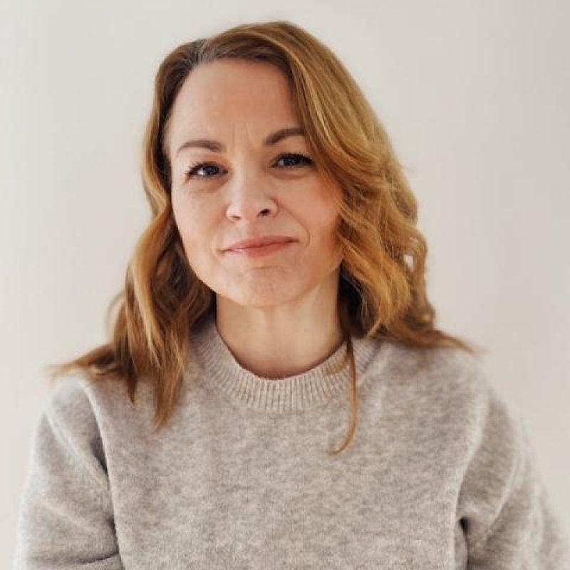 A woman with blonde hair wearing a grey sweater sits in front of a white wall