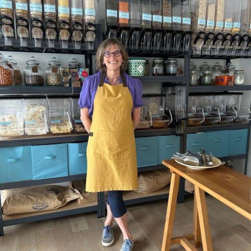 A smiling woman wearing a yellow apron stands in front of shelves holding jars of grains and cooking ingredients