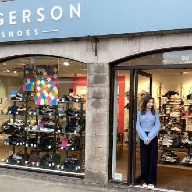A woman wearing a blue shirt stands in a doorway next to windows displaying several pairs of shoes at Rogerson Shoes in Perth