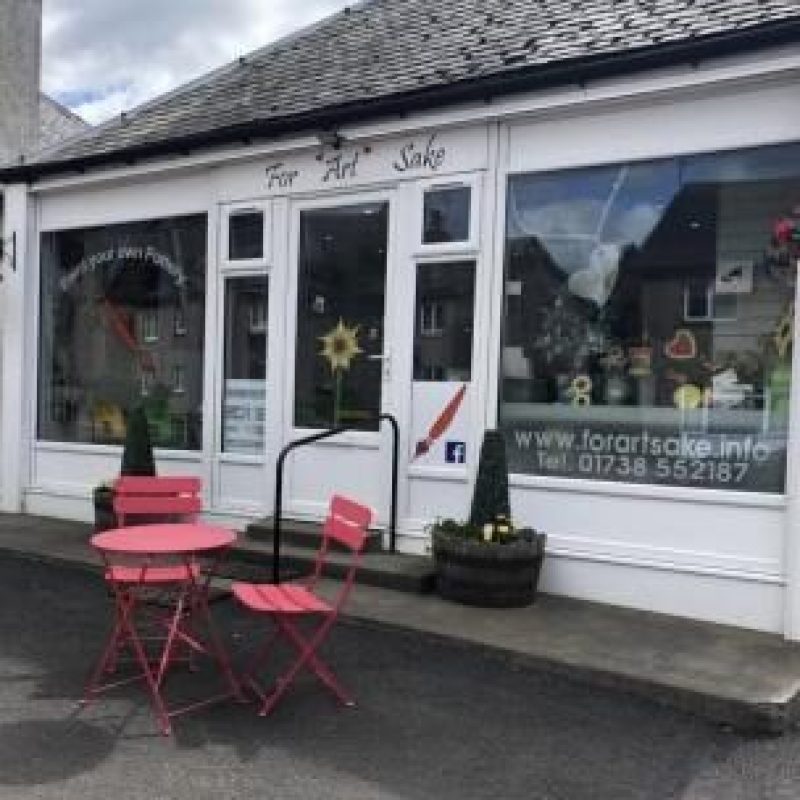 A small white building with large windows has a sign reading For Arts Sake written in cursive, and sits next to outdoor chairs