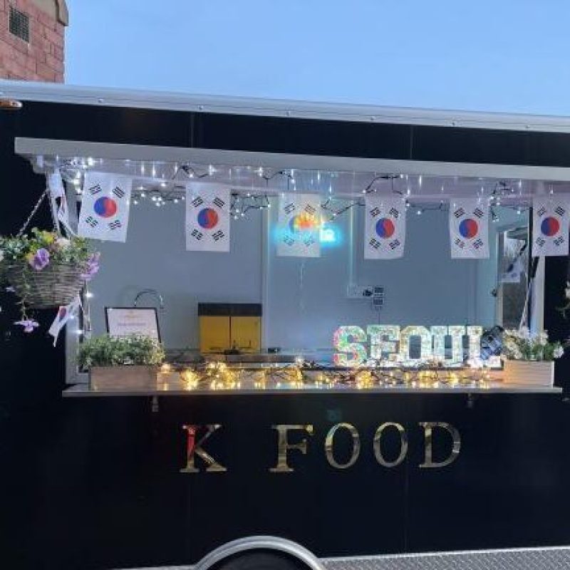 A black food truck with a serving window decorated with fairy lights, South Korean flags, and gold lettering reading K Food