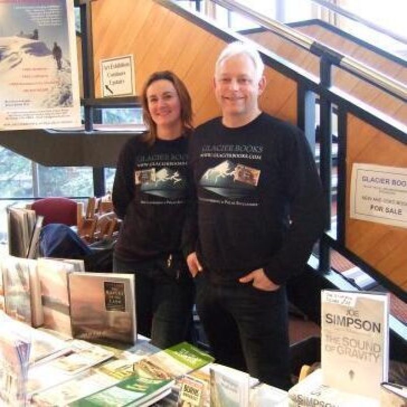 A smiling man and woman wearing matching black printed shirts stand next to each other behind a stall table covered in books