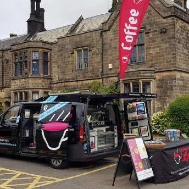 A black van decorated with a white mug is full of barista supplies and a coffee machine and is parked in front of a service table