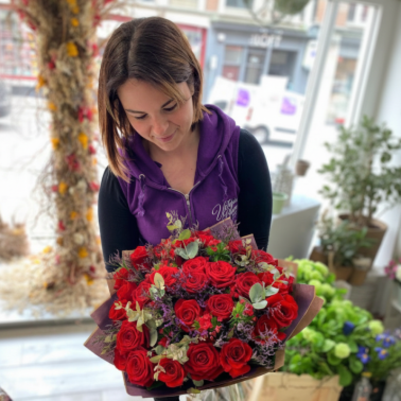 A woman wearing a purple jumper looks down at a bouquet of red roses with green leaves that she is holding