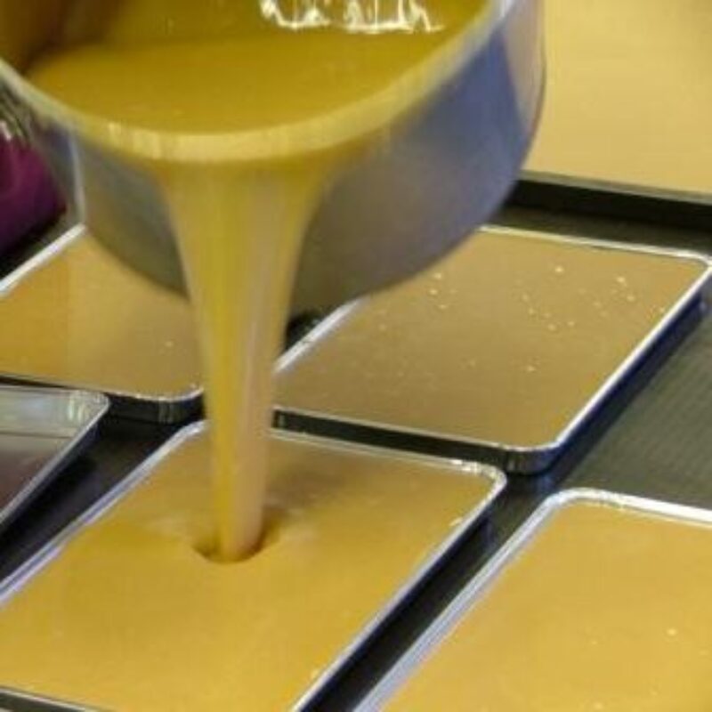 Thick yellow liquid is poured from a pot into shallow rectangular trays arranged in rows