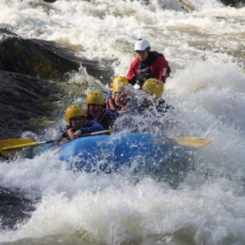 A group of people wearing wet suits and yellow helmets hold ores and sit in a raft going through large river rapids