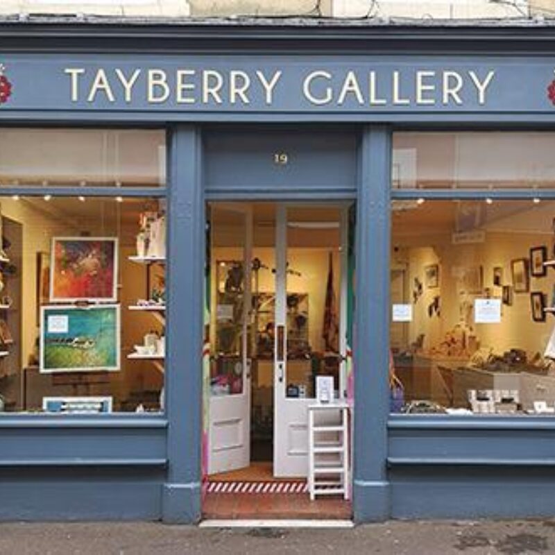 A grey shop has two large windows displaying hanging artwork and paintings on shelves either side of a doorway
