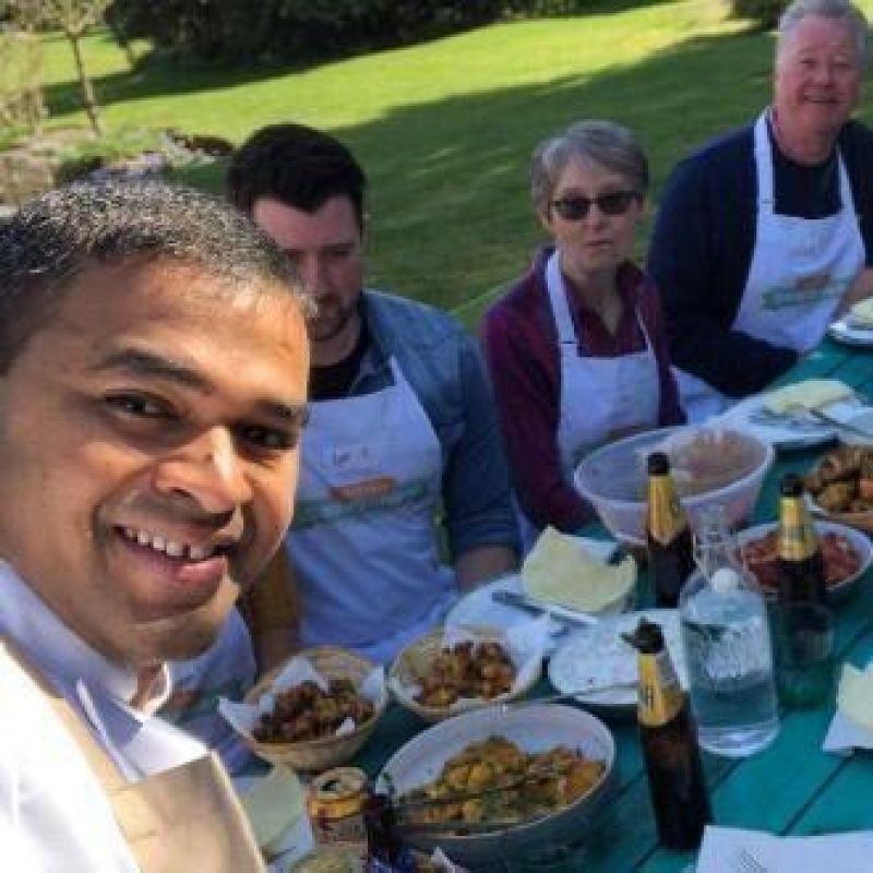 A group of people wearing matching white aprons sit around an outside bench covered in a spread of an array of food and wine